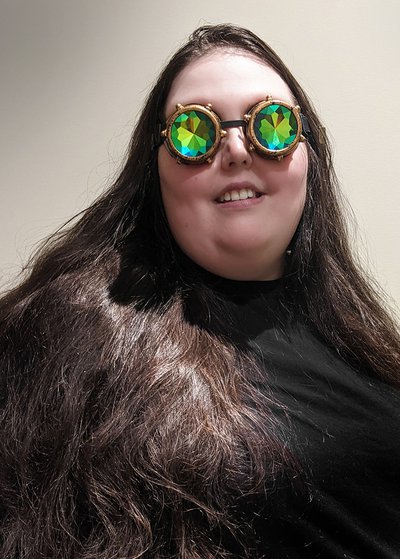 Susan, a pākehā person with long brown hair and steampunk goggles, smiles at the camera