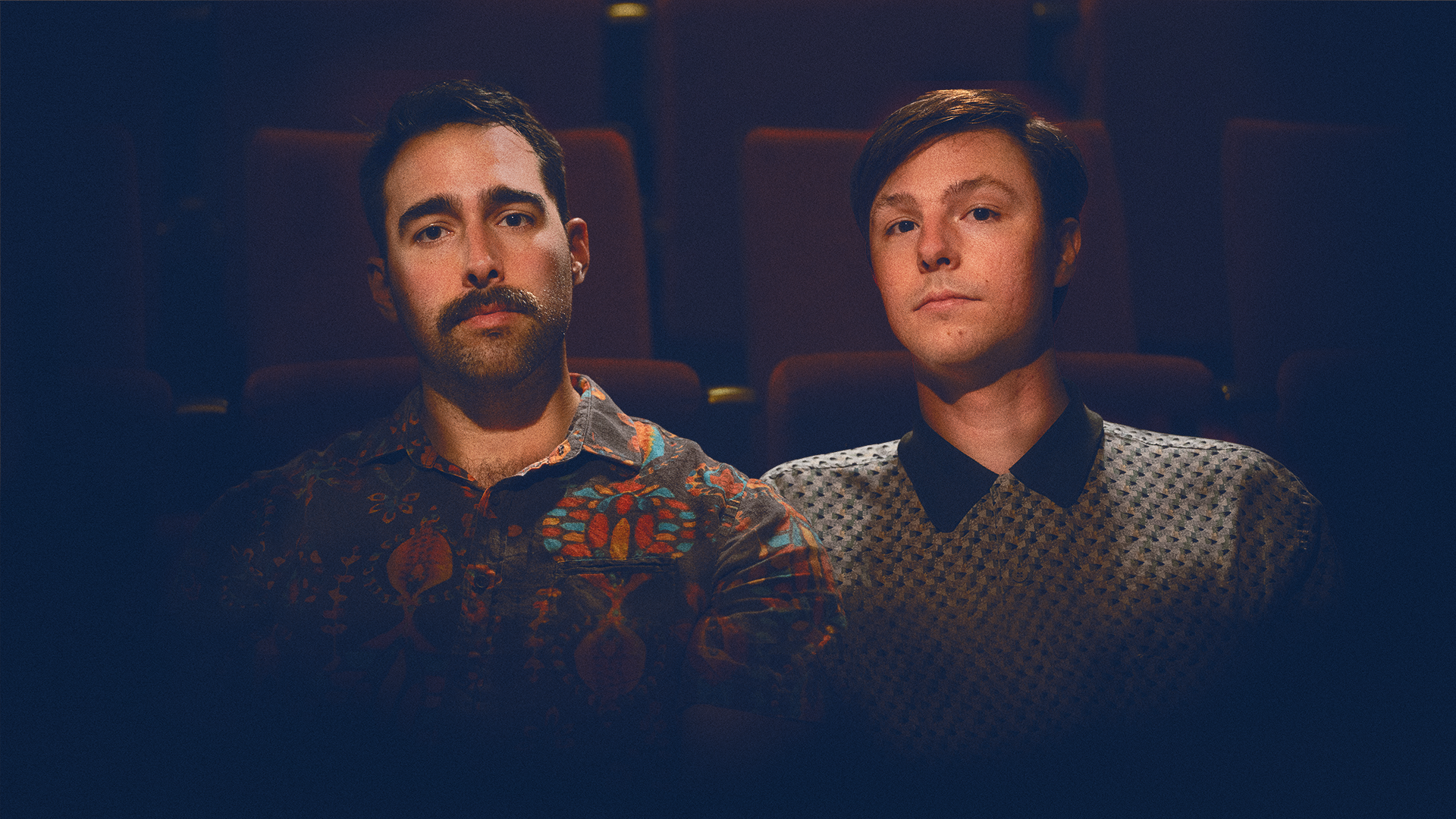Max and Ben sit together in a darkly lit theatre. The two are  close and face the viewer..png