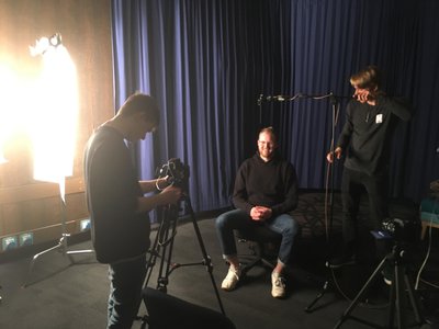 TABS - An individual being filmed in front of a curtain with someone behind the camera and someone operating a boom mic